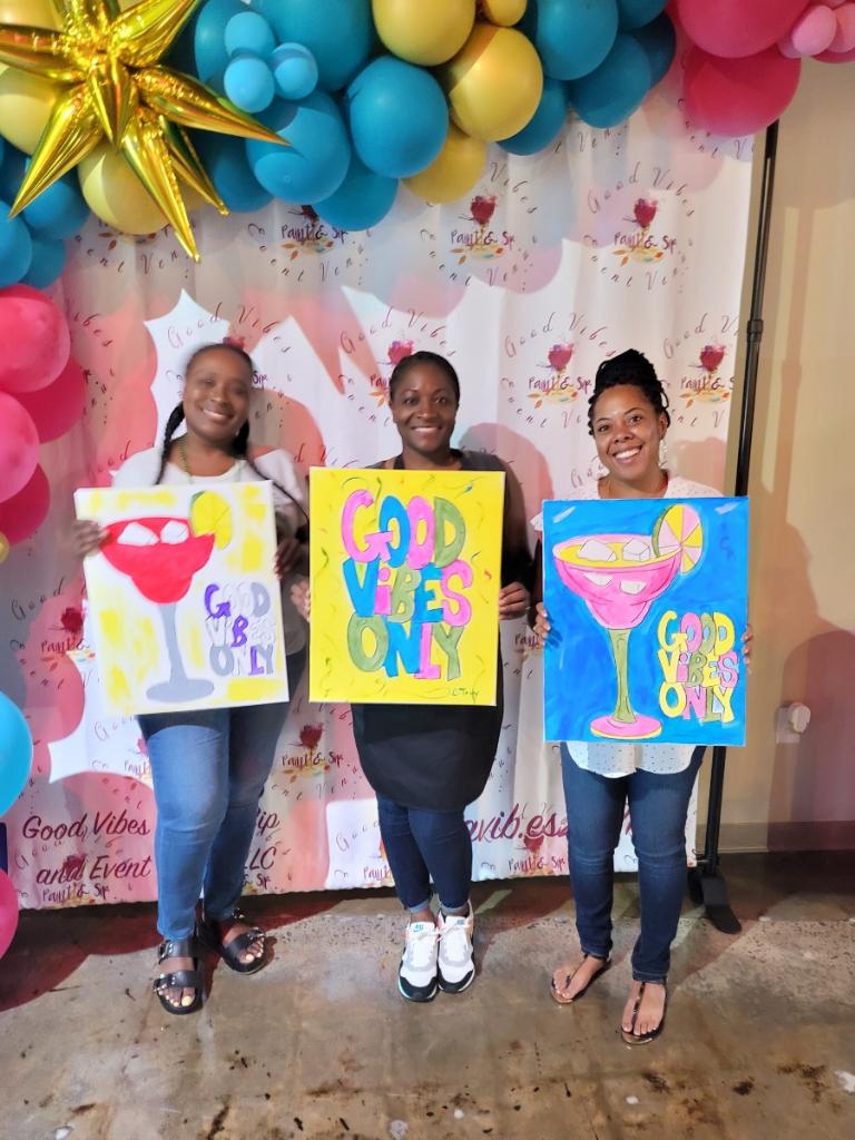 Three women holding up paintings of a drink and a heart.
